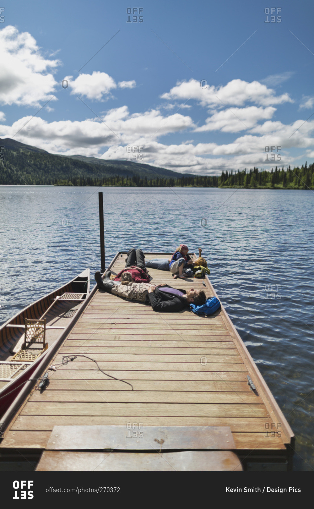 Three adults and a young girl lounging on a boat dock on a lake soaking up the sun, Byers Lake campground, Denali State Park, Alaska, United States of America