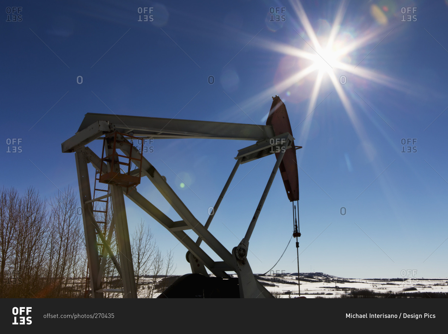 Pumpjack and snow covered hills with sun burst and blue sky, Longview, Alberta, Canada