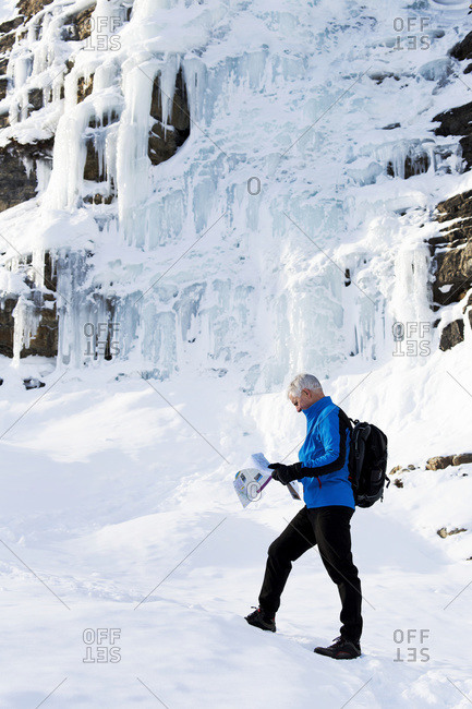 Male hiker on snow covered trail looking at map with a rock cliff covered in ice, Lake Louise, Alberta, Canada