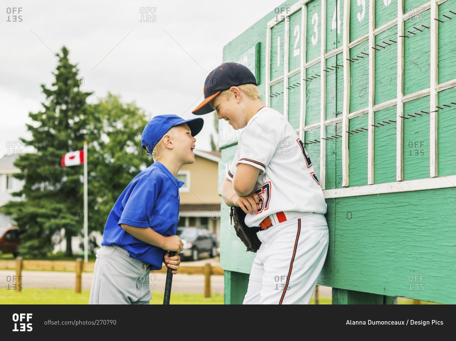 Two boys in baseball uniforms playfully argue in front of the scoreboard during a baseball game at a sports field, Fort McMurray, Alberta, Canada