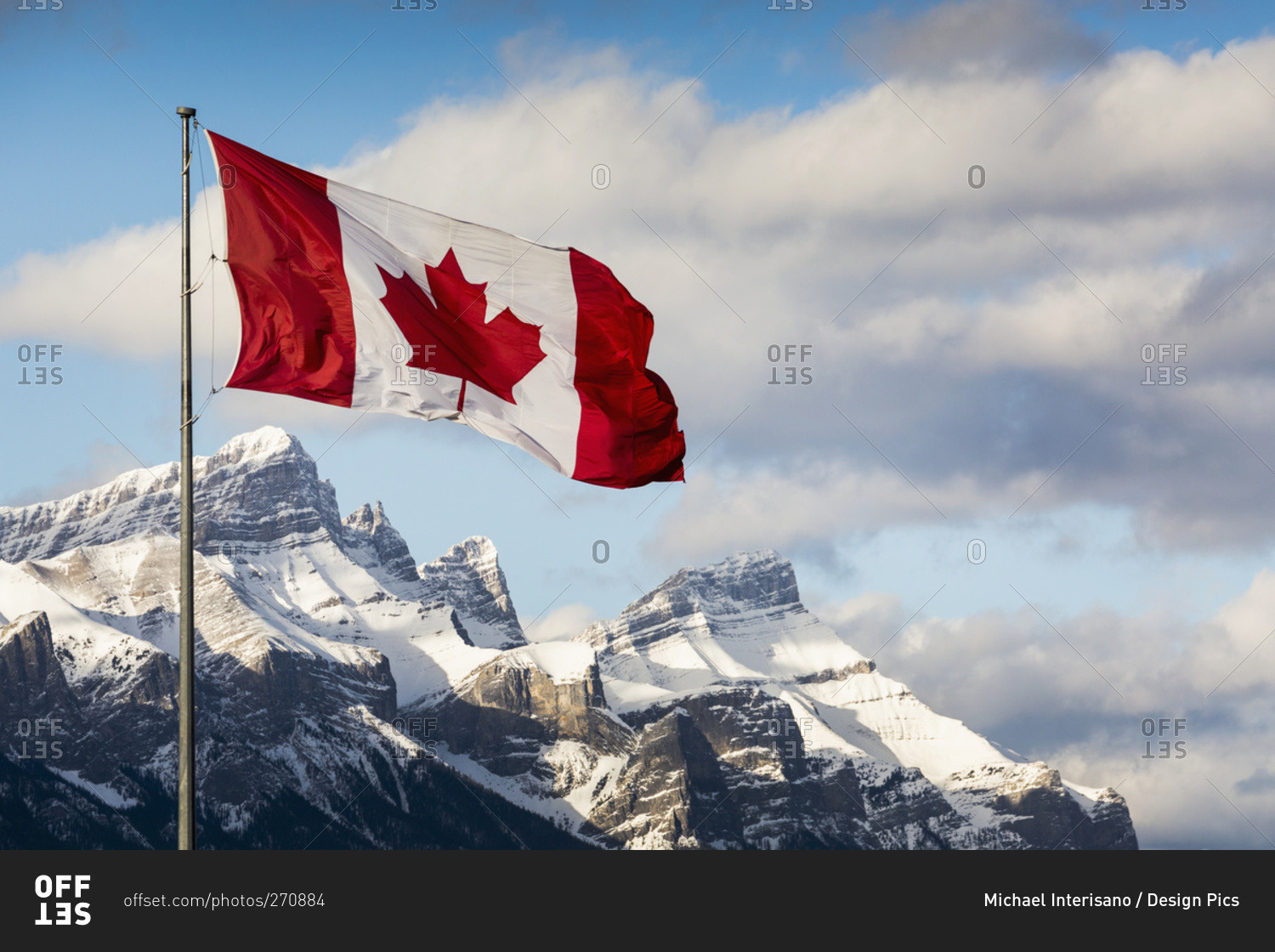 Canadian flag blowing in the wind on a flag pole with snow covered mountain range in the background with blue sky and clouds, Canmore, Alberta, Canada