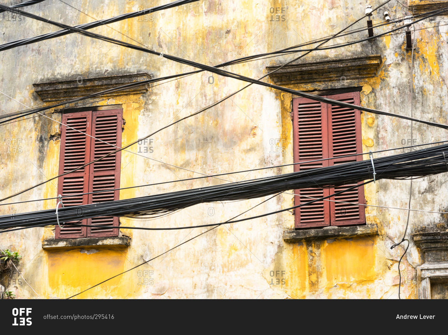 An old yellow building with two windows with brown shutters with a chaotic collection of cables in the foreground in the city of Hanoi, Vietnam