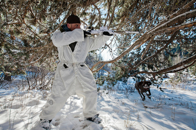 A hunter wearing a cold-weather camouflage outfit aims his rifle in preparation to shoot while his hunting dog prepares to retrieve