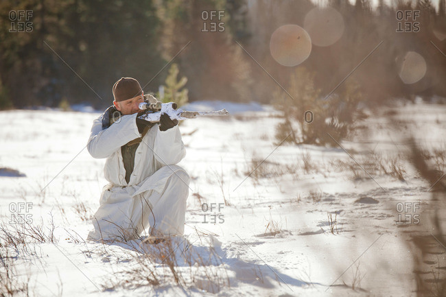 A hunter wearing a cold-weather camouflage outfit aims a rifle while crouching in a snow-covered field