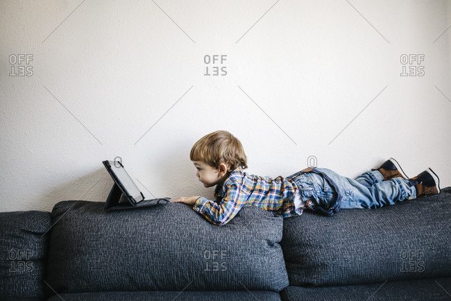 Little Boy Lying On Backrest Of A Couch Looking At Digital Tablet