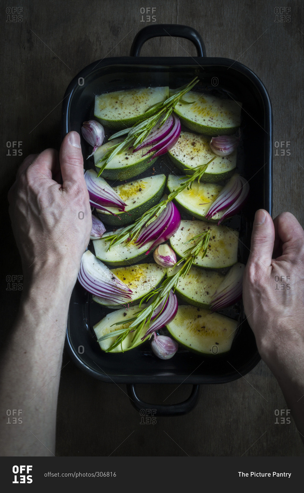 Hands holding an oven dish with courgette, onions and fresh herbs prepared for cooking