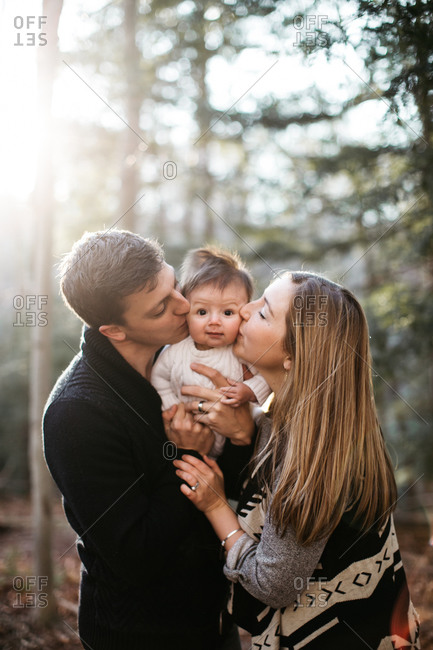 Cute baby with spiky hair and bemused expression being kissed by his parents  in woods stock photo - OFFSET