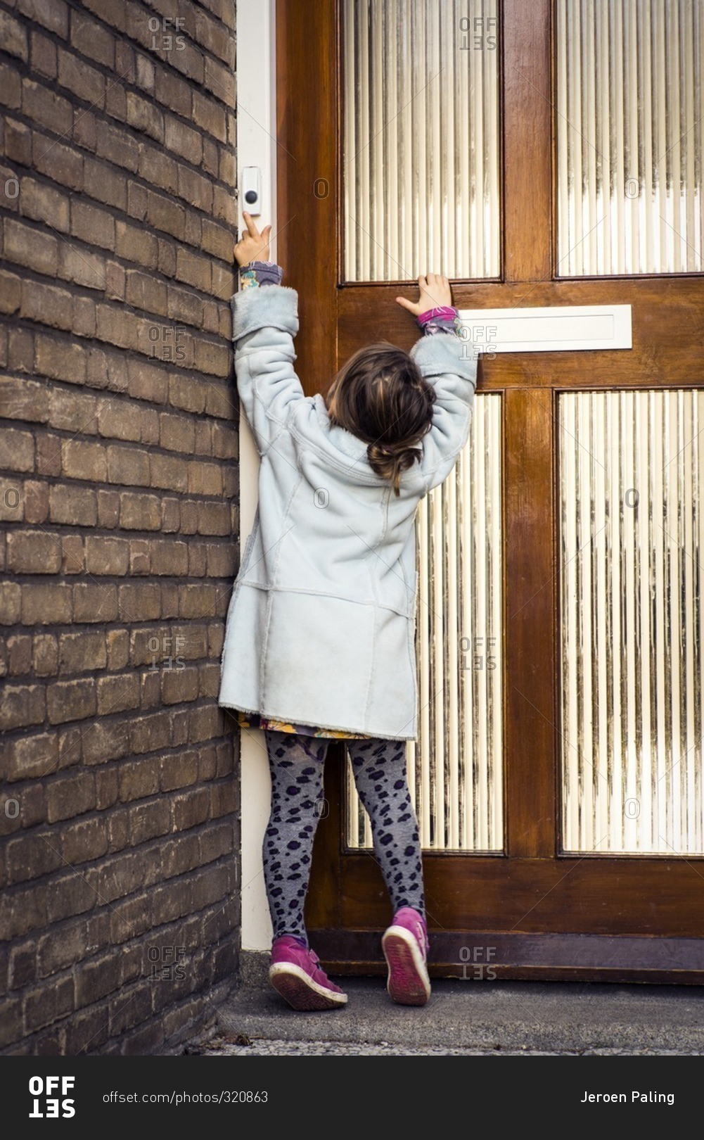 Little girl stands on tiptoe at door to ring doorbell stock photo - OFFSET