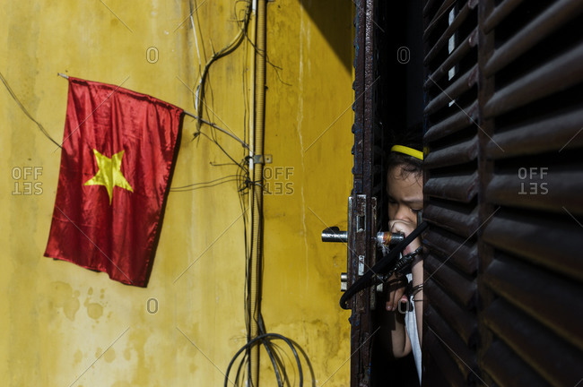 Young girl peaking through a heavily chained door next to a yellow wall emblazoned with the flag of Vietnam