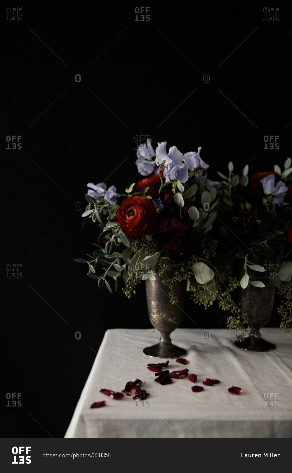 Still life with vases holding roses and purple flowers beside rose petals on a white tablecloth