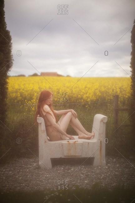Woman sitting on a stone bench in a sheer dress looking out over a field of colza flower