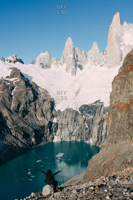 Person with arms raised sitting on a boulder looking out at a snow covered mountain peak rising behind a clear glacial lake