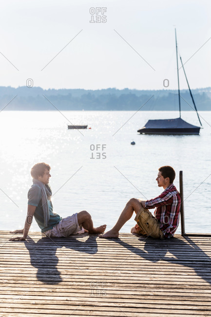 Friends sitting face to face on wood pier next to lake, Schondorf, Ammersee, Bavaria, Germany