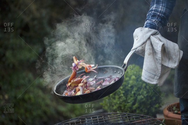 A man holding a frying pan sauteing vegetables above an open fire
