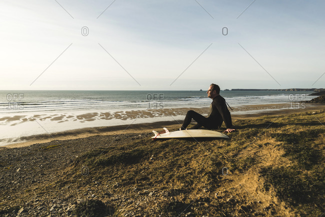France, Bretagne, Finistere, Crozon peninsula, man sitting at the coast with surfboard