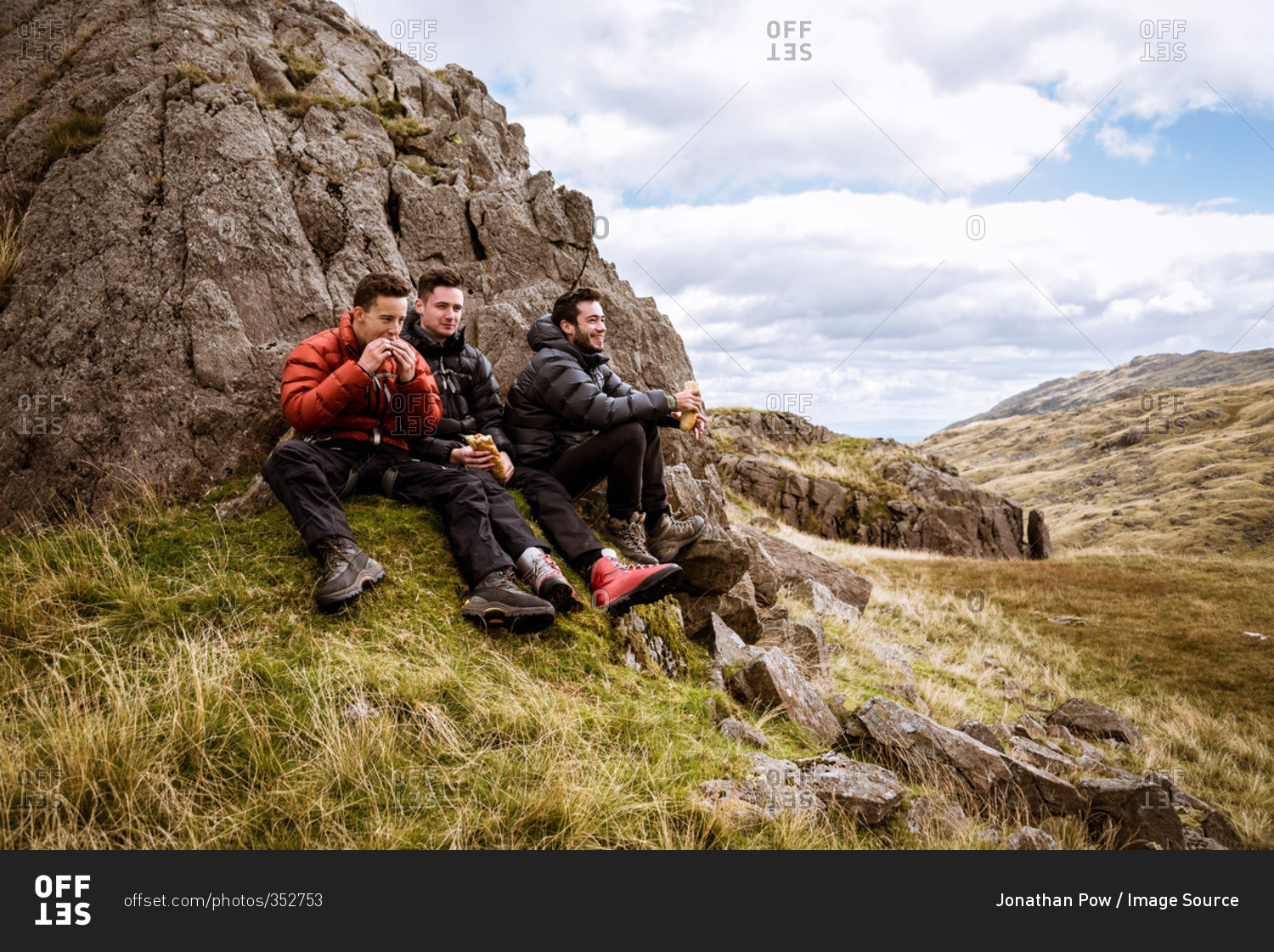 Three young male hiking friends eating sandwiches, The Lake District, Cumbria, UK