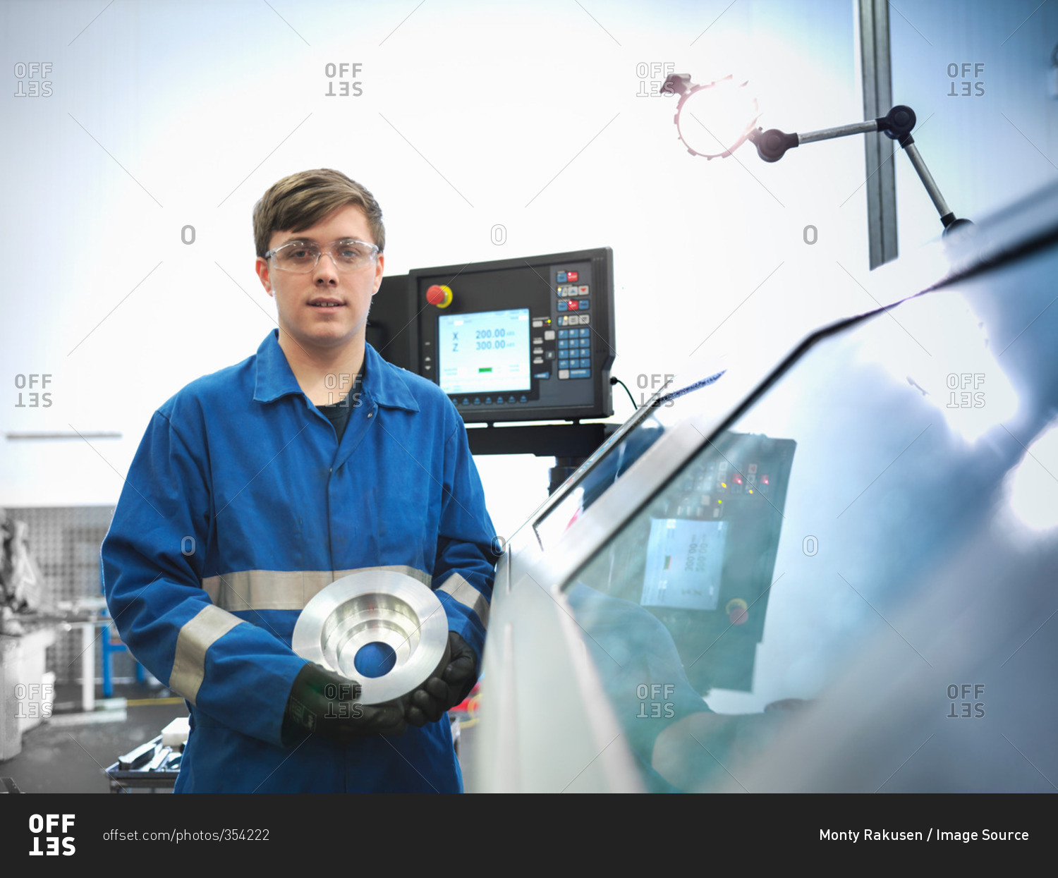 Portrait of apprentice engineer holding steel component with lathe in engineering factory