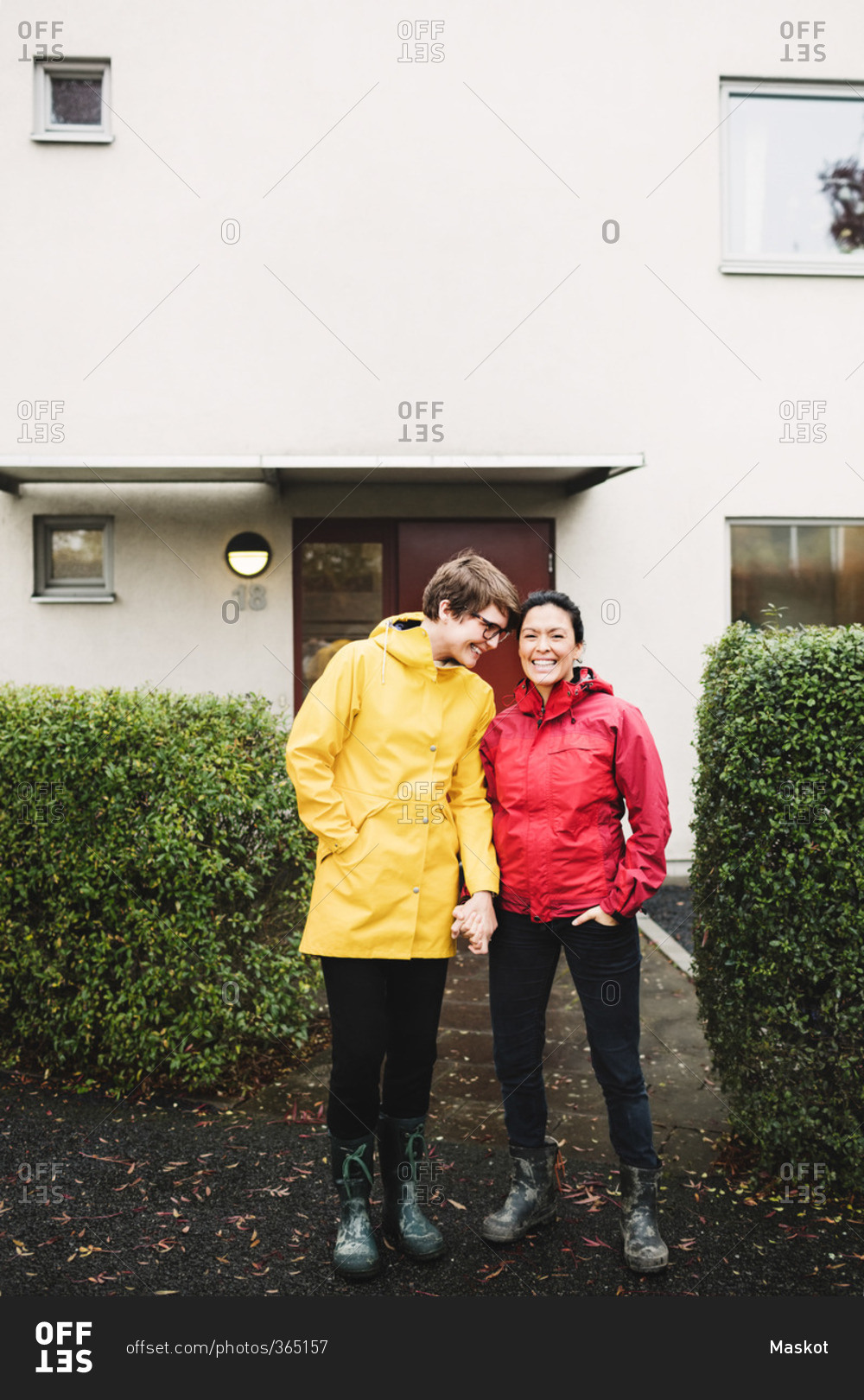 Smiling lesbian couple wearing raincoat standing outside house