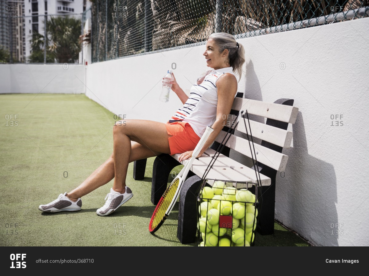 Smiling mature woman holding water bottle while sitting on bench at tennis court