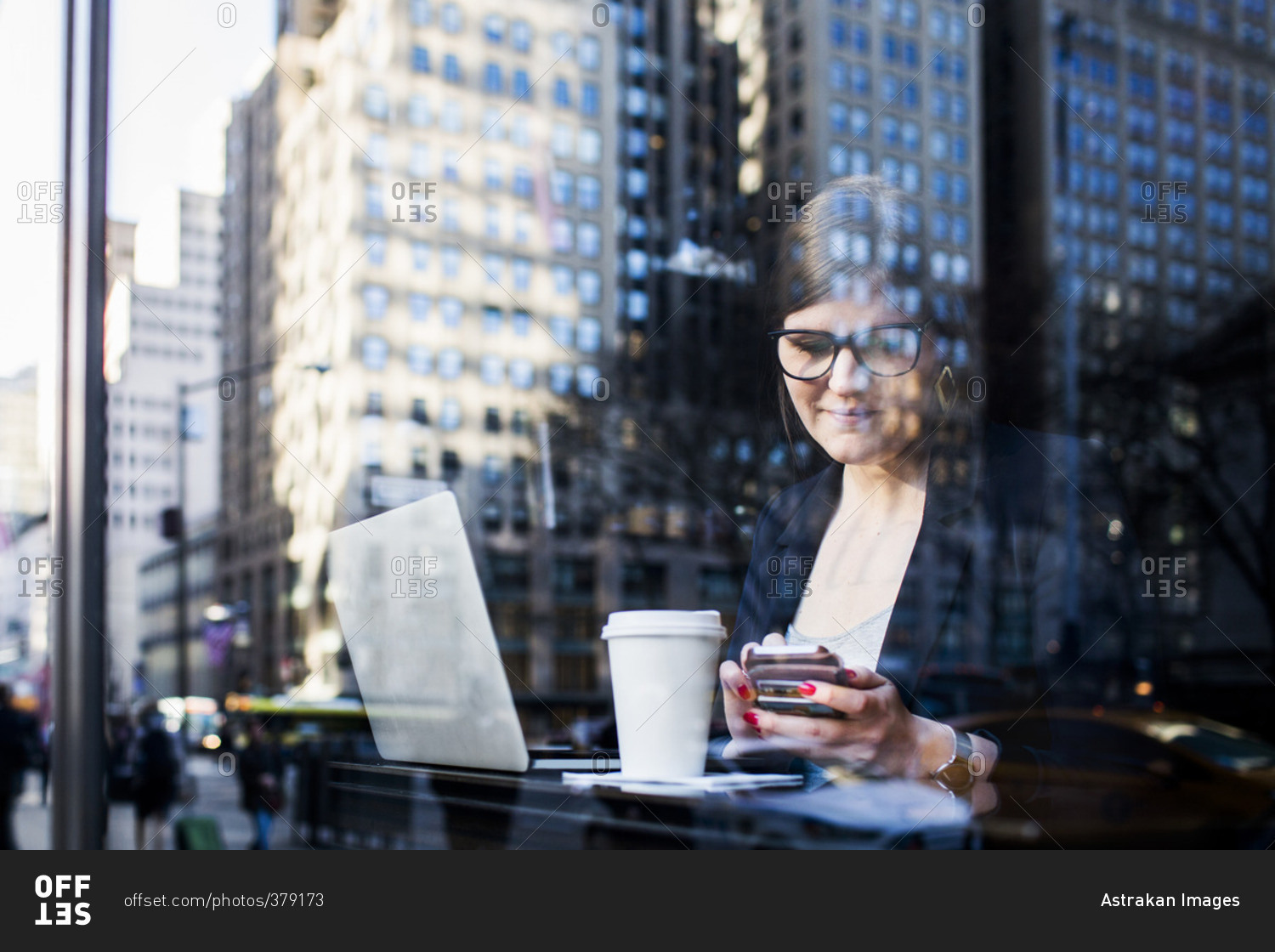 Businesswoman with technologies at cafe seen through window reflecting buildings