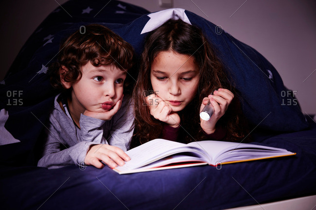 Brother and sister under duvet reading book by torchlight