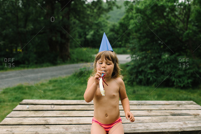 Girl sitting on a picnic table wearing a birthday hat and blowing a party noisemaker