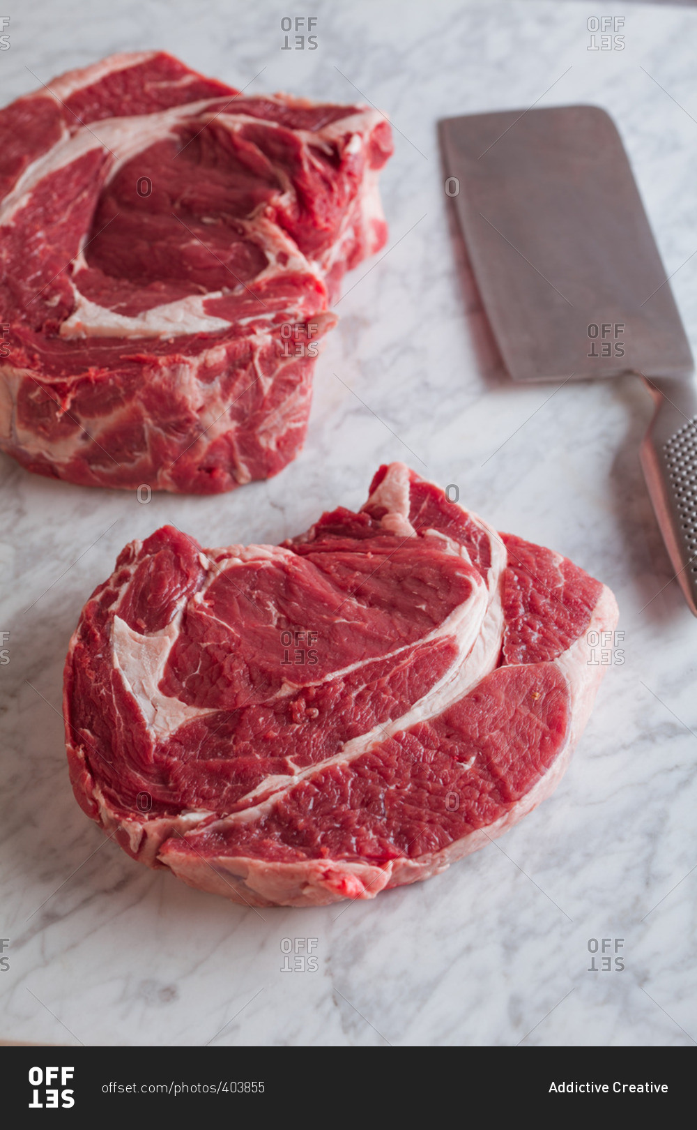 Raw thick steaks and a meat cleaver on a marble surface stock photo