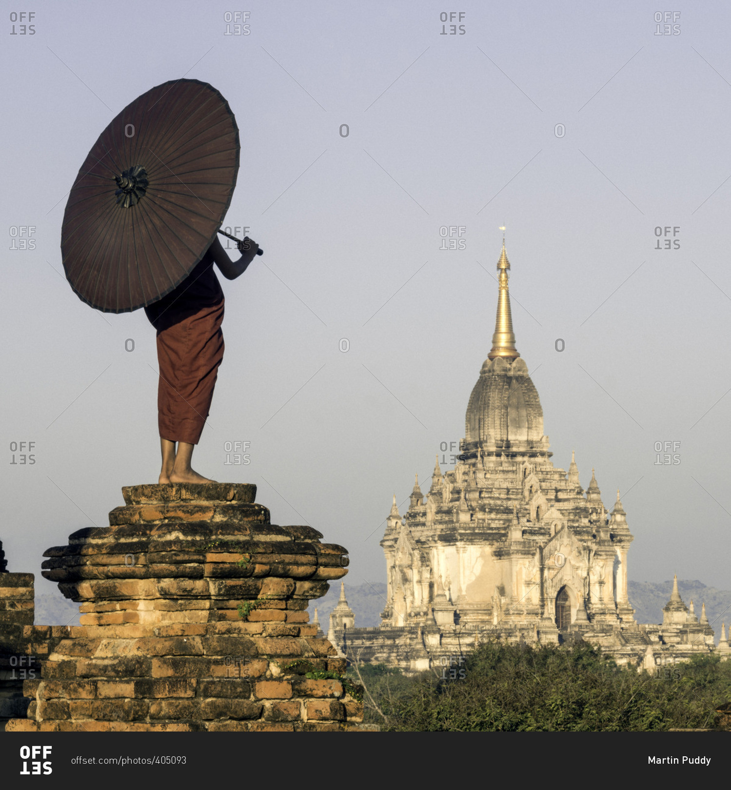 Young Buddhist monk standing on temple top holding a parasol in front of an ancient temple