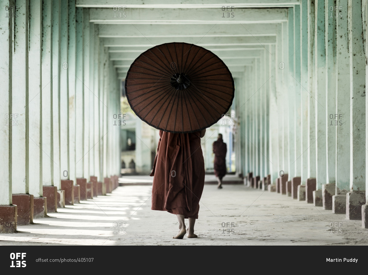 Young novice Buddhist monk walking along a temple complex corridor using a parasol