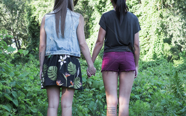 Back view of two fashionable female friends standing hand in hand, partial view