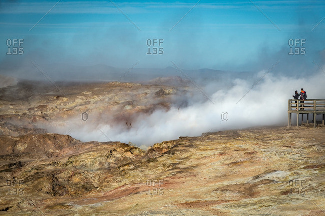 Two people on boardwalk overlooking Icelandic geothermal site with steam rising
