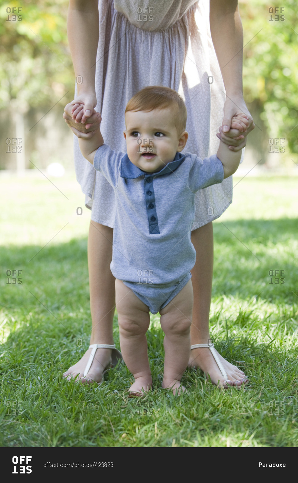 Baby standing outdoors, holding mother's hands for support