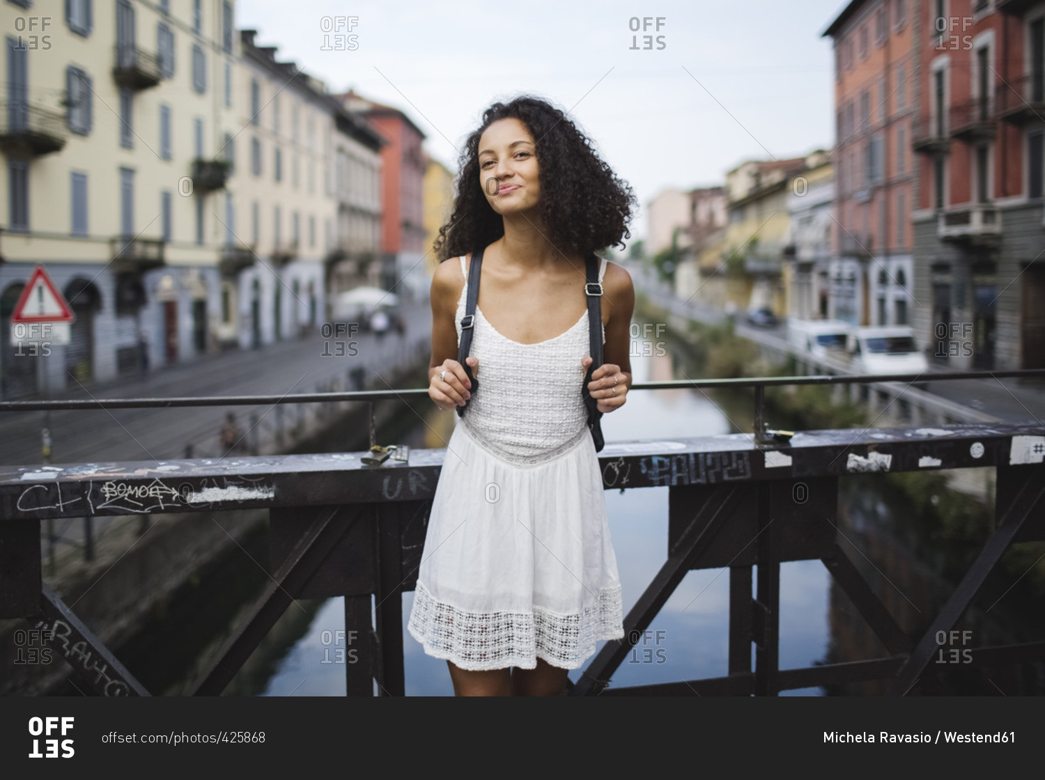 Italy- Milan- portrait of smiling young woman with backpack wearing white summer dress standing on a bridge