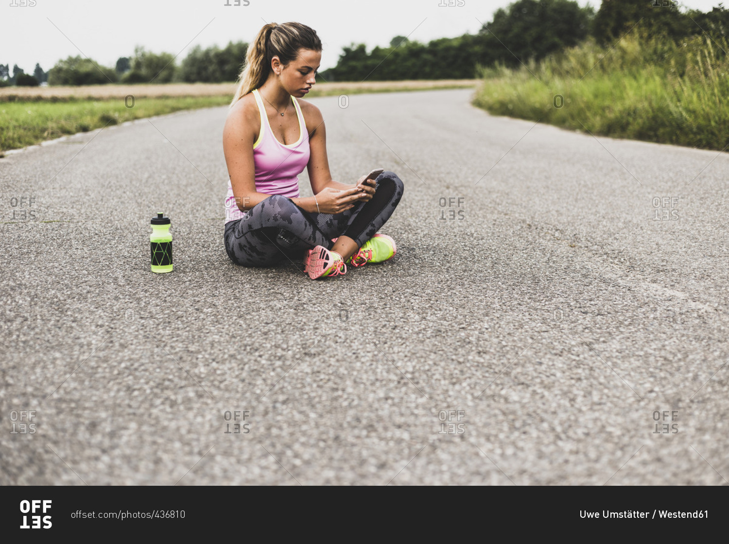 Sportive young woman sitting on street with cell phone and drinking bottle