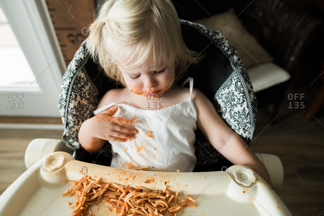Girl sitting in a high chair eating a messy meal of spaghetti noodles and tomato sauce