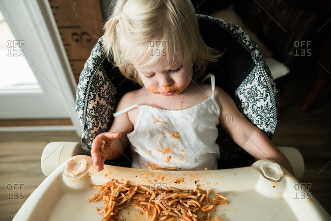 Girl sitting in a high chair eating a messy meal of spaghetti noodles