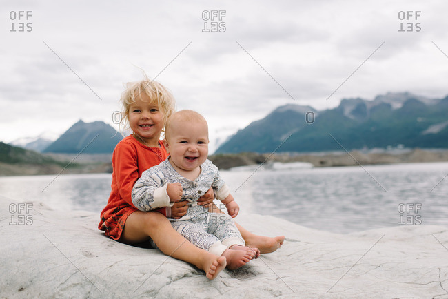 Two young children sitting together on a large rock overlooking a lake