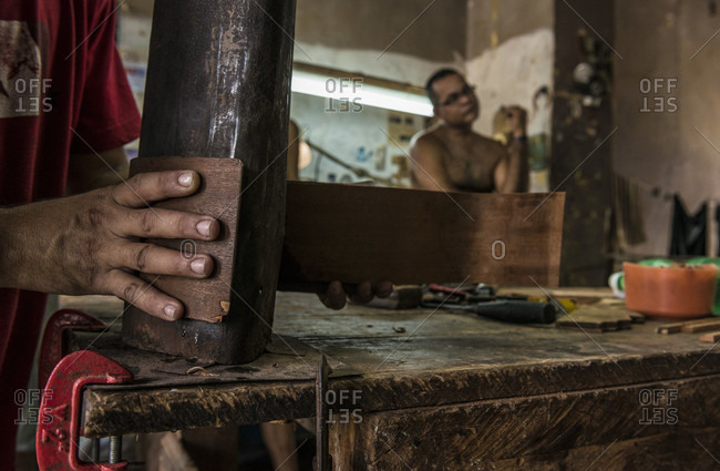 Havana, La Habana, Cuba - April 29, 2014: Close up of the hand of a young Cuban guitar-maker bending wood to shape with a hot coal-fired bending jig in a hand-made guitar workshop in Havana, La Habana, Cuba. One hand presses the wet wood against the form, and the other coaxes it into shape. His co-worker waits in the background