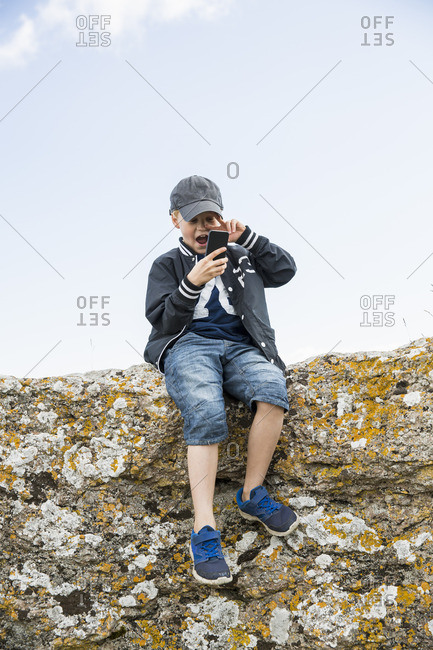 Sweden, Gotland, Boy sitting at edge of cliff and using telephone