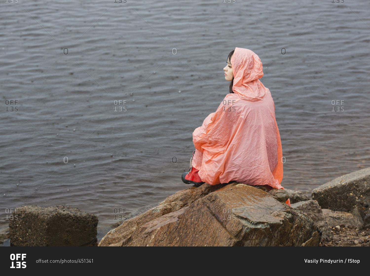 Thoughtful woman wearing raincoat sitting on rock at lakeshore during rainy season
