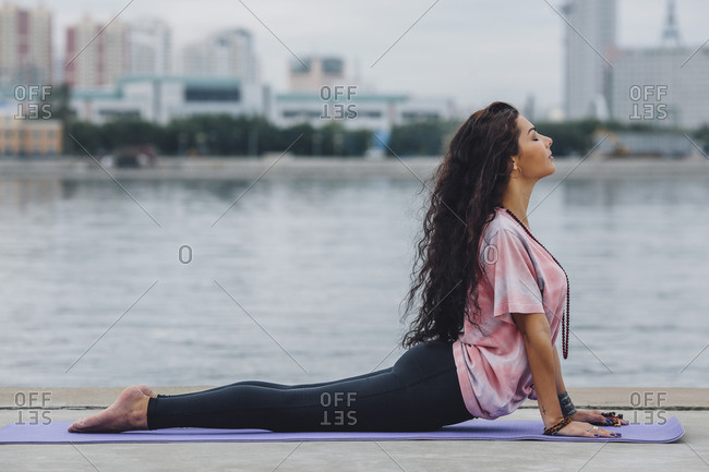 Active young woman exercising Cobra pose on yoga mat near river