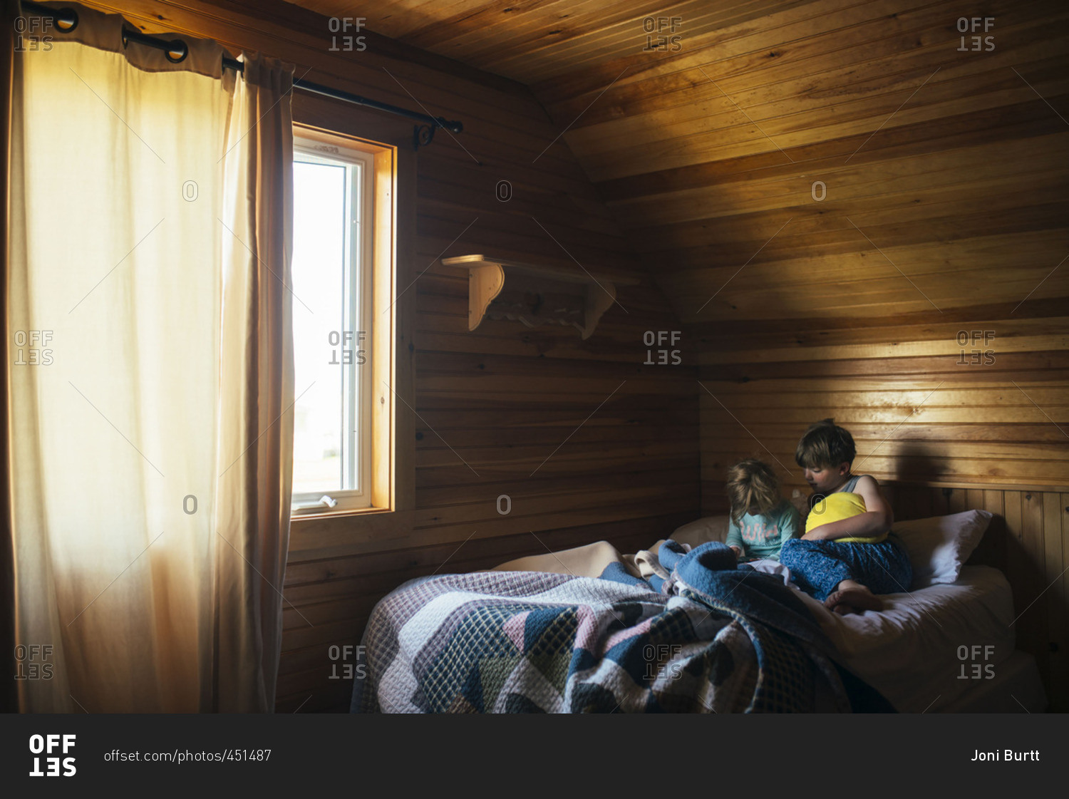 Brother And Sister Sitting Together On Bed In Room With Wood Paneling