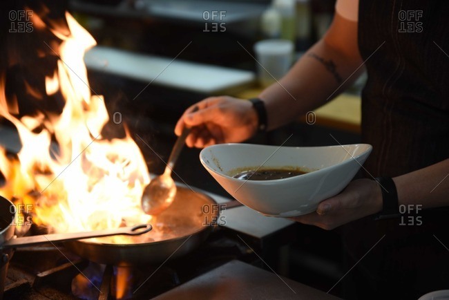 Person adding a spoonful of liquid to a flaming pan on a stove