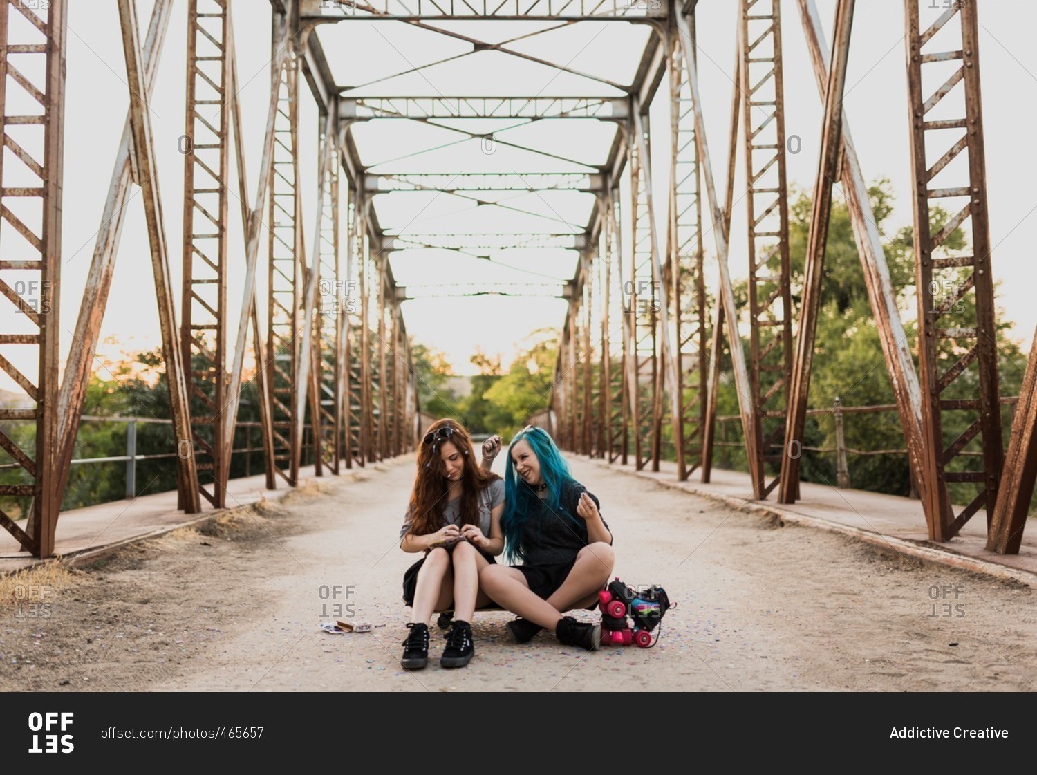 Two teen punk girls sitting on skateboard on a bridge rolling a cigarette