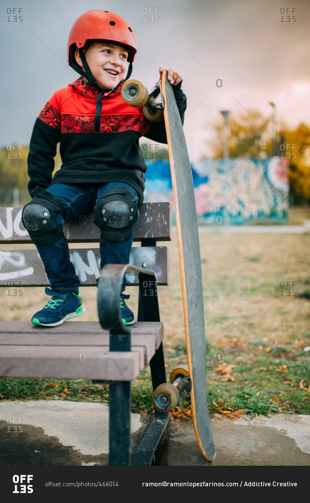 Smiling kid with red helmet and knee pads with long board on a bench at the skate park