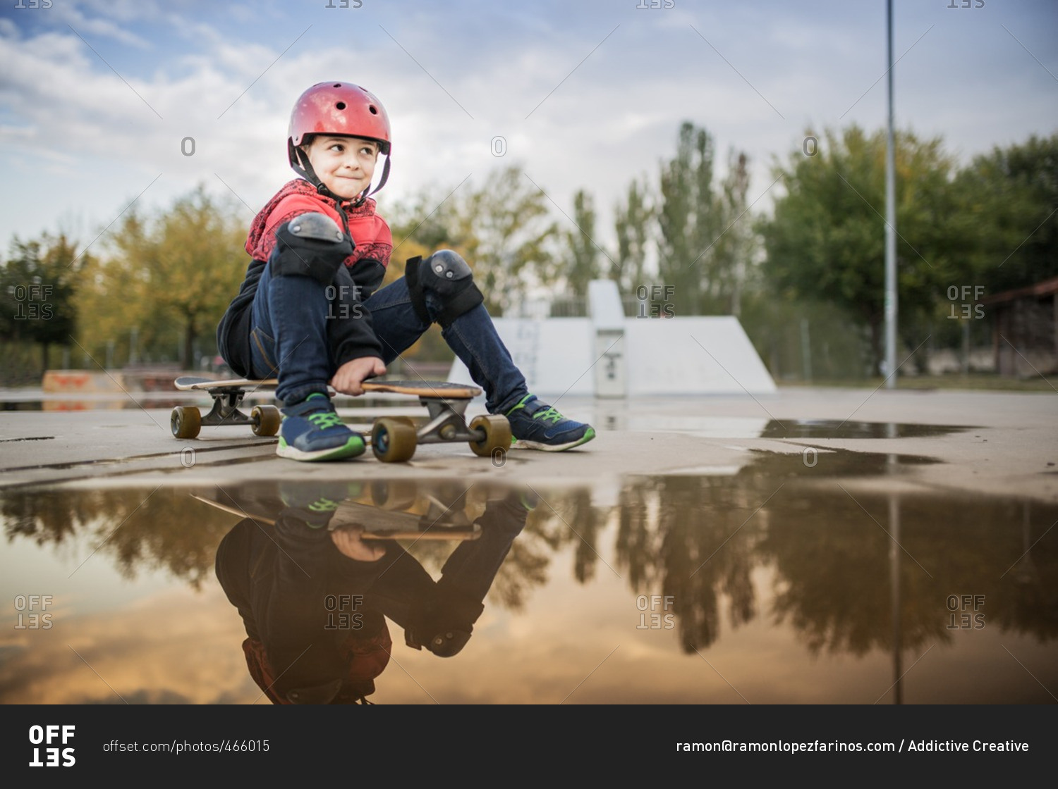 Smiling kid with red helmet and knee pads with long board by a puddle at the skate park