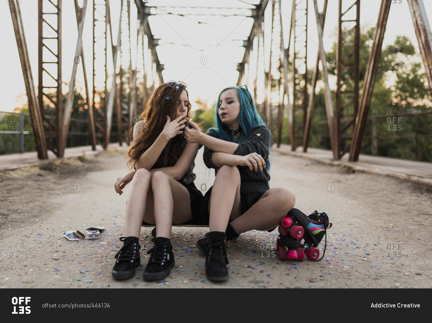 Teen punk girl sitting on skateboard on a bridge lighting her friend's cigarette