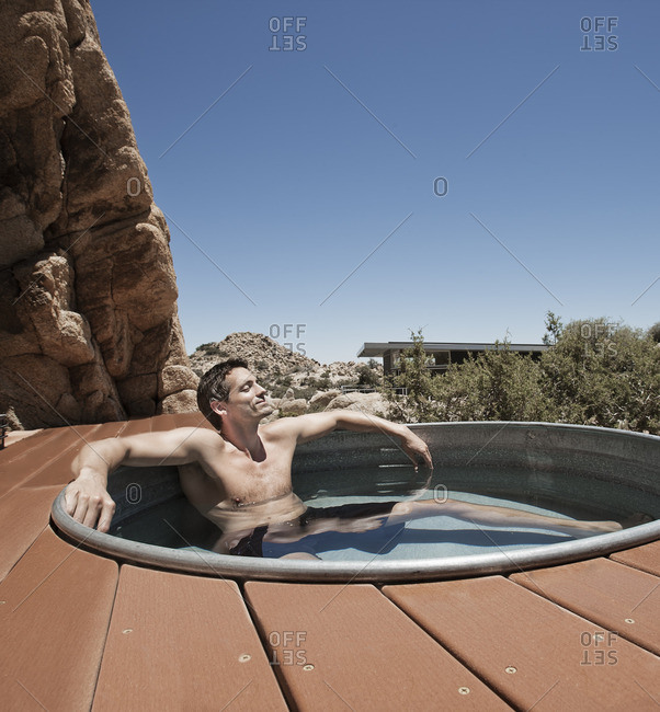 A man on the terrace of an eco home, a low impact house in the desert landscape, in a sunken hot tub