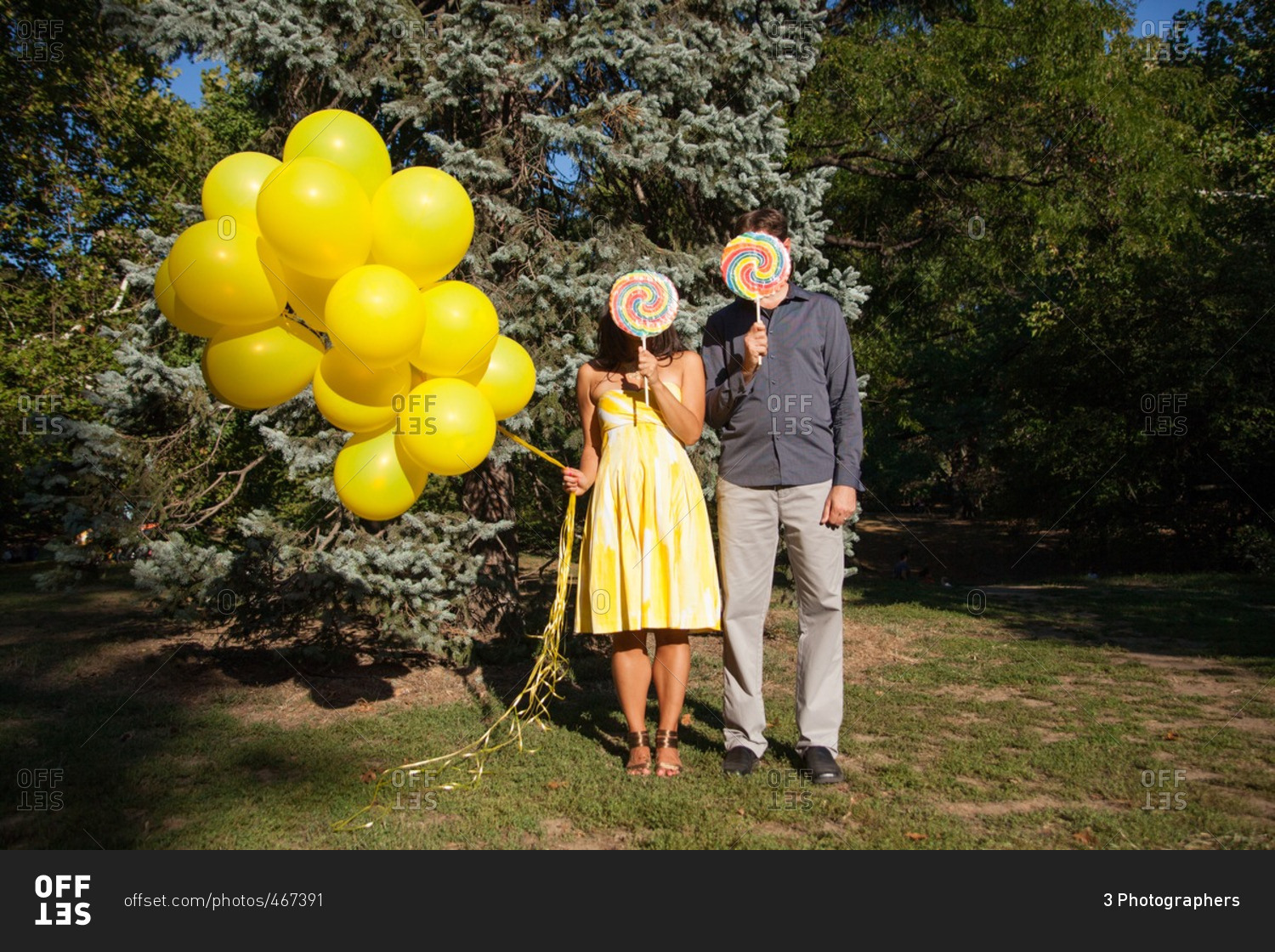 Couple standing in Central Park holding lollipops in front of their faces and yellow balloons