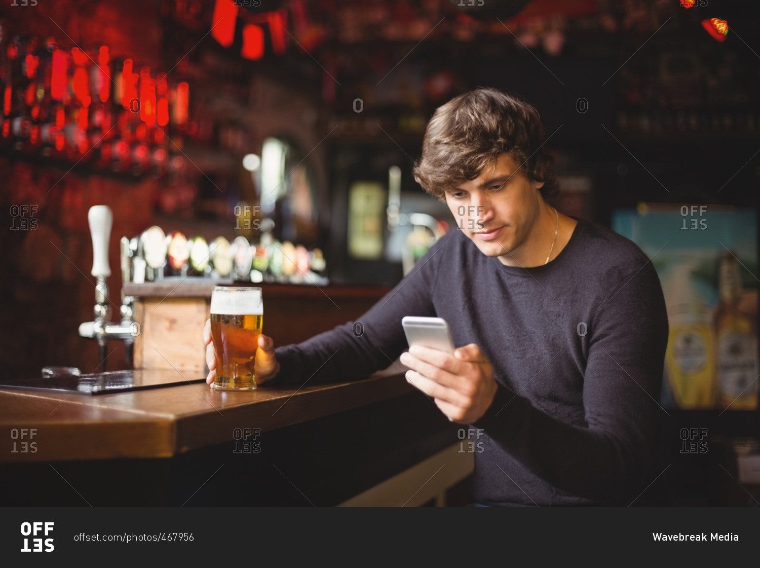 Man using mobile phone with glass of beer in hand at bar